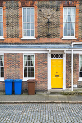 Abstract city street scene in England with a yellow door on a brick town house and trash containers in front of the building. 