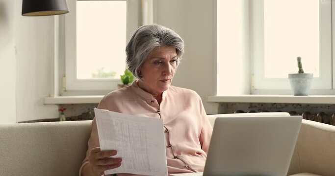Thoughtful old woman accountant working online from home review paper documents bills check conditions terms of payments on laptop. Serious older Latina lady think on rental tenancy payments using pc