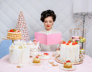 Beautiful brunette woman reads a magazine with a pink cover on the background of cakes and pies and various confectionery