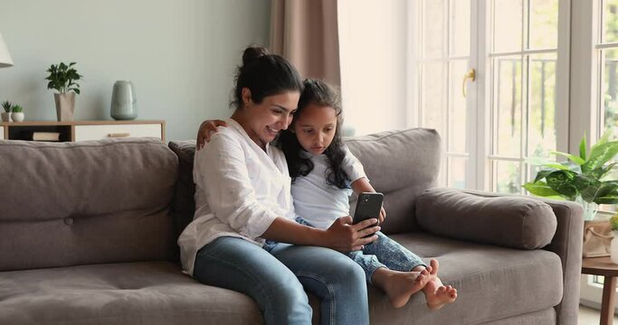Two Indian sisters grownup older younger kid best friends sit on couch hug check social media news make video call using mobile phone. Bonding mommy little daughter watch funny pictures on cell online