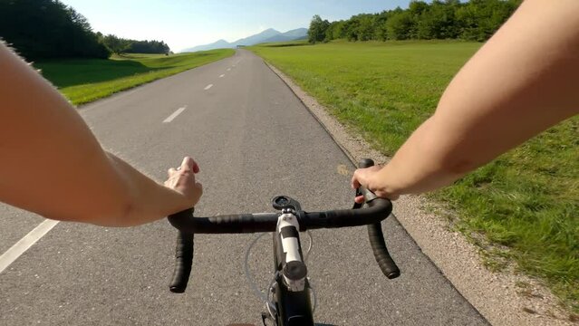 Caucasian female racing cyclist riding on a paved road through the green nature and looking at mountain landscape in front, point of view shot.