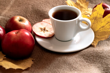 Cup of herbal tea with fresh red apples surrounded with yellow leaves on woolen textile surface