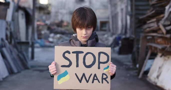 Ukrainian little sad angree boy shows card Stop War standing on destroyed street, child refugee, protest against war, russian war in Ukraine, invasion, humanitarian crisis, victim of military conflict