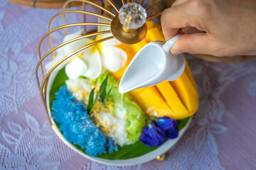 Mango sticky rice mixed with coconut milk on a banana leaf with white ceramic dish on wooden table and white clothing cover is a popular traditional dessert in Thailand.
