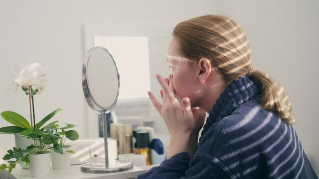 a woman smears her face with cream at home sitting in front of a mirror. a shadow falls on her from the blinds.