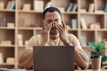Tired arab freelancer guy sitting at workdesk in front of laptop at home and rubbing his eyes,...