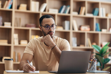 Focused arab man in glasses using laptop sitting at desk and writing in notebook, watching webinar...