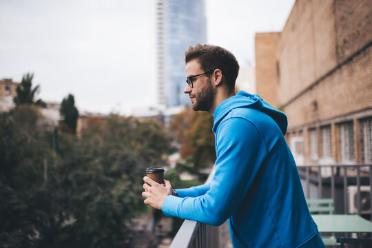 Side view of Caucasian male model with disposable takeaway cup of caffeine beverage thinking about youthful lifestyle, millennial hipster guy with coffee to go thoughtful looking away at terrace
