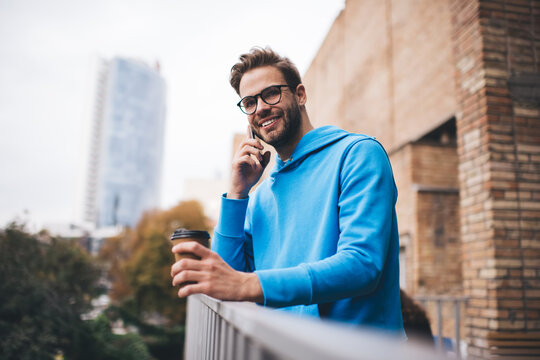 Cheerful hipster guy in optical spectacles calling to oculist for discussing vision protection and correction, happy male in eyeglasses millennial user with coffee to go phoning via mobile application