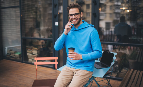 Half length of happy millennial man in classic eyewear calling to best friend for communicate during coffee break in city, cheerful hipster guy with takeaway cup using roaming connection for phoning