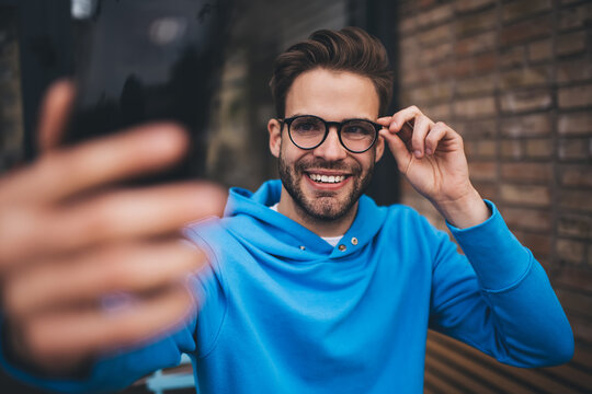 Smiling hipster guy shooting video vlog via application on mobile technology, funny millennial male blogger connecting to 4g on smartphone gadget for clicking selfie photos during leisure in city