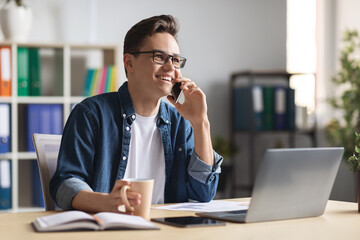 Fototapeta premium Smiling millennial businessman talking on cellphone and drinking coffee at workplace in office