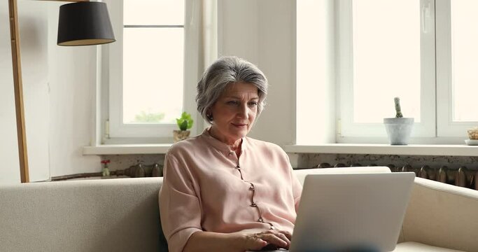 Older Hispanic woman confident laptop user engaged in freelance work from home browse internet type document sitting on cozy couch at living room. Middle aged retired lady learn online using computer