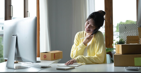 A portrait of Asian woman, e-commerce employee sitting in the office full of packages on the table using a laptop and smartphone, for SME business, e-commerce, technology and delivery business.