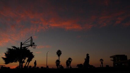 People playing basket ball game, silhouettes of players on basketball court outdoor, sunset ocean beach, California coast, Mission beach, USA. Black hoop, net and backboard on streetball sport field.