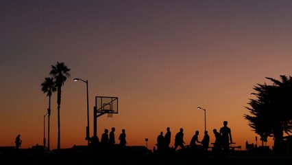 Fototapeta na wymiar People playing basket ball game, silhouettes of players on basketball court outdoor, sunset ocean beach, California coast, Mission beach, USA. Black hoop, net and backboard on streetball sport field.