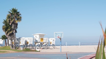 Palm trees and basketball sport field or court on beach, California coast, USA. Streetball playground on shore and lifeguard stand, tower ot station. Mission beach, San Diego. Hoop, backboard and sky.