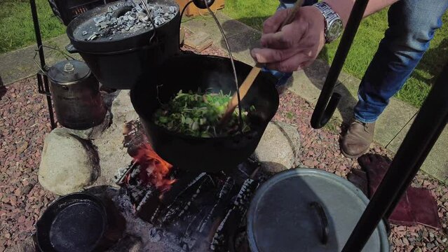 Cooking outside in the garden on a campfire, unrecognizable man Stir in the cast iron pan with fresh vegetables. low angle view cooking outdoors food preparation.