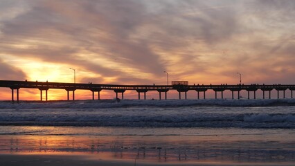 Obraz premium Silhouette of people walking, surfers surfing by pier in sea water. Ocean waves, dramatic sky at sunset. California coast, beach or shore vibes at sundown. Summer seascape seamless looped cinemagraph.