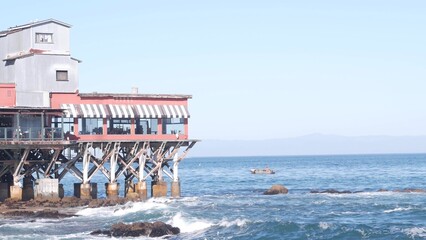 Fototapeta na wymiar Waterfront beachfront cafe on piles, pillars or pylons, Monterey beach, California coast aesthetic, USA. Sea ocean water waves and building architecture. Summer tourist vacations in watersibe resort.