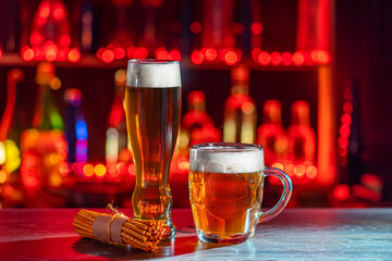 Glass of beer on the bar counter. Jugs, mugs, pints of brew beverage, ale, cider on the wooden table in pub, bar. Backlit dark room, showcase with craft beer bottles in the brewery