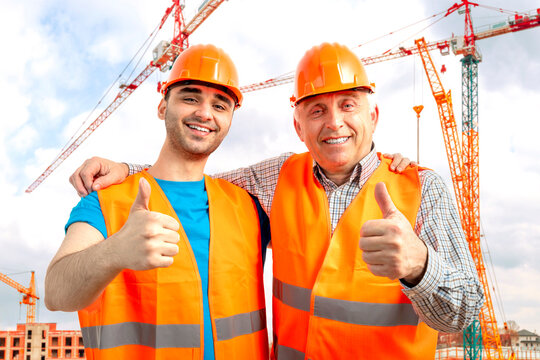 Older Construction Supervisor With Younger Worker Helper At Construction Site. Managers Wearing Protective Workwear, Hard Hat, Construction Cranes On Skyline. Construction Workforce, Working Labor Man