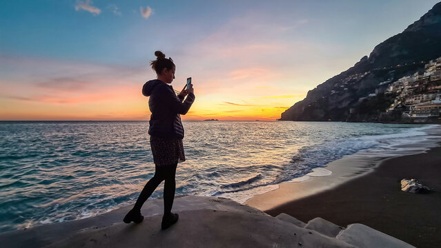 Woman taking pictures while watching sunset on Marina Grande Beach and colorful buildings of hillside village Positano, Amalfi Coast, Italy, Campania, Europe. Vacation at Tyrrhenian, Mediterranean Sea