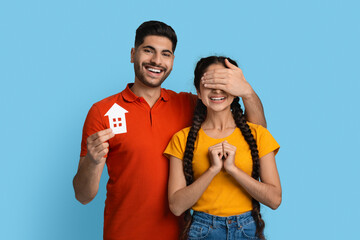 Happy arab guy holding paper house icon and covering girlfriend's eyes