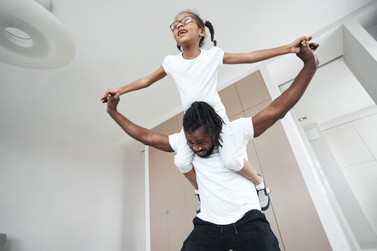 Afro American girl sits on her father's neck with her arms wide apart