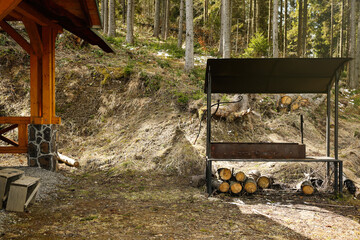 Metal barbecue with stacked firewood and wooden gazebo near forest