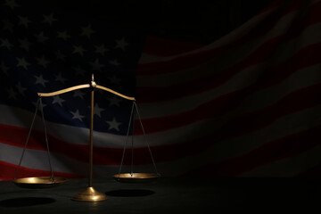 Scales of justice on black table against American flag in darkness, space for text