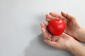 Elderly woman holding red heart in hands on light grey background, top view. Space for text