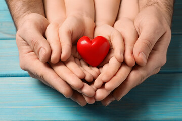Parents and kid holding red heart in hands at light blue wooden table, closeup