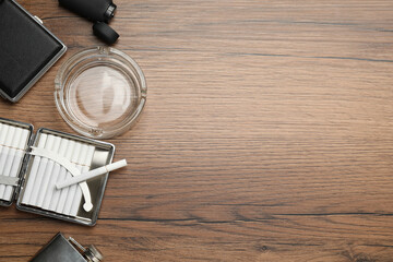 Open case with tobacco filter cigarettes, glass ashtray, lighter and pocket flask on wooden table, flat lay. Space for text