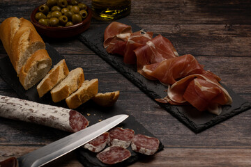 food concept: view of two slate stone plates with cold meats, ham, salami, bread, olive oil, olives, on a wooden table. iberian cured meats.  