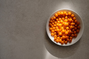 Organic sea buckthorn in a white plate. Layout on a gray table, flat lay. Fresh orange berries in bright sunlight. Natural valuable food supplement.