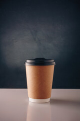 Brown paper disposable cup of coffee, cappuccino with plastic cap on white table, dark background. Tea to go. Copy space, vertical shot