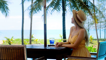 Fototapeta na wymiar Tropical vacation. Smiling happy woman in straw hat sitting in outdoor seaside cafe, drinking refreshment drink and enjoy fresh air and beautiful view of sea and palm tree. Summer holidays