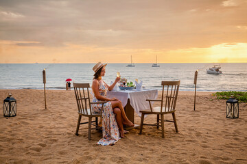 Romantic date on the sunset beach. Woman sitting alone on the table with gourmet food, drinking champagne from glass. Happy woman in luxury outdoor restaurant happy and smiling after proposal