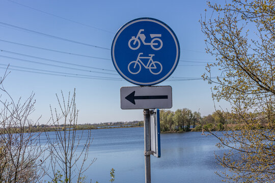 Traffic sign indicating: lane for bicycles and mopeds with a black arrow pointing the direction, blue and white round plate, water of a river in the background in the Netherlands