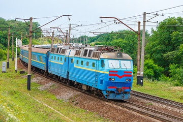 Naklejka premium A powerful two-section blue locomotive pulls a long passenger train to the railway station. Trees with green leaves. Evening summer lighting.