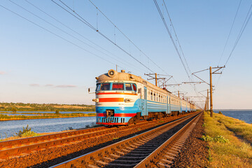 Fototapeta na wymiar A multi-car commuter passenger electric train travels along a straight road on the shore of a large lake. Sunset summer lighting.
