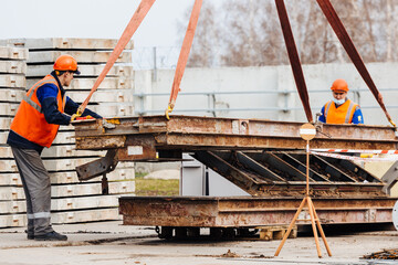 Fototapeta premium Slinger in helmet and vest controls unloading of metal structures on construction site. White handyman unloads load.