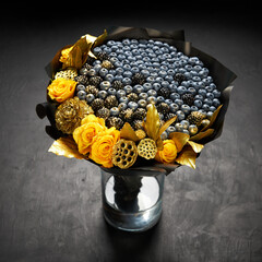 Cool bouquet of blueberries and blackberries decorated with yellow roses and gold leaves standing in a vase on a black background