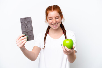 Young reddish woman isolated on white background taking a chocolate tablet in one hand and an apple...