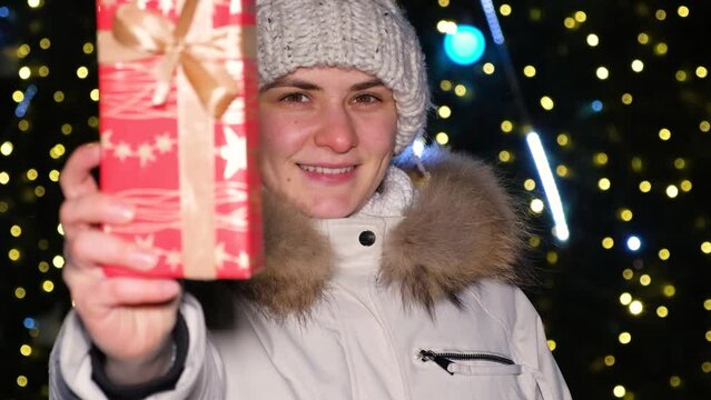 A happy woman shows a red gift box and looks into the camera standing near the Christmas tree
