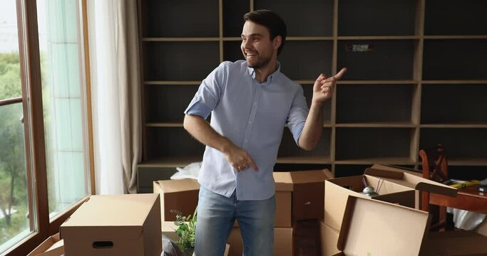 In unfurnished living room with heap of carton boxes with stuff, carefree guy celebrate relocation day to new first affordable dwelling, listen music dance looks happy. Bank loan, rental flat concept