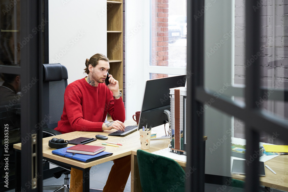Poster Serious young manager having a conversation on mobile phone while working on computer at his workplace at office