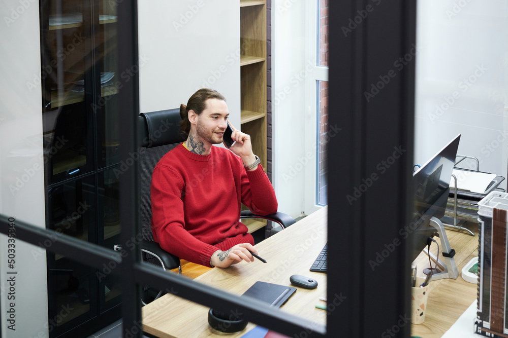 Poster Smiling manager sitting at table at office in front of computer monitor and having business conversation on mobile phone