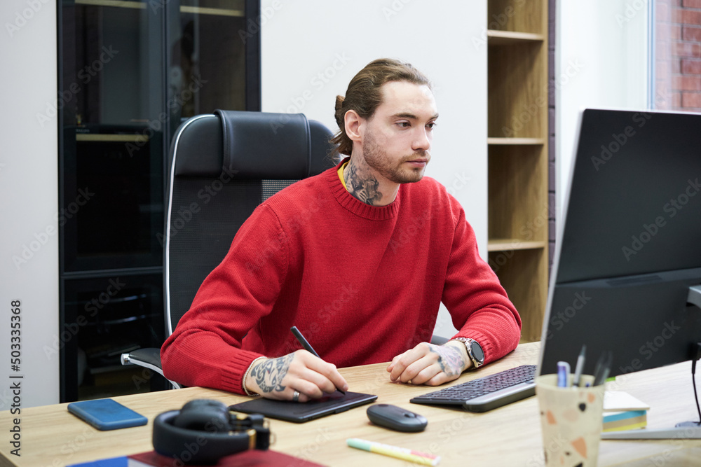 Aufkleber Young designer sitting at desk in front of computer monitor and using graphic tablet to do his work